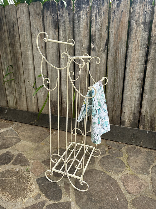 Decorative metal stand with a wooden fence and stone ground in the background. Metal Towel Rail w Shelf Small Aged Cream