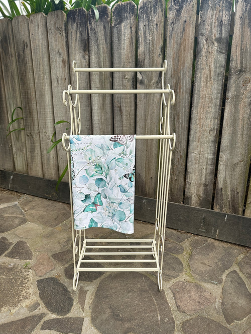 White metal clothes rack with a towel on it, against a wooden fence background. Metal Towel Rail w Shelf Small Aged Cream