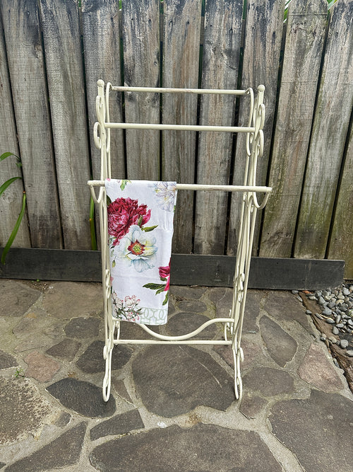 White metal towel rack with a floral towel against a wooden fence background. Metal Filigree Small Towel Rail Aged Cream