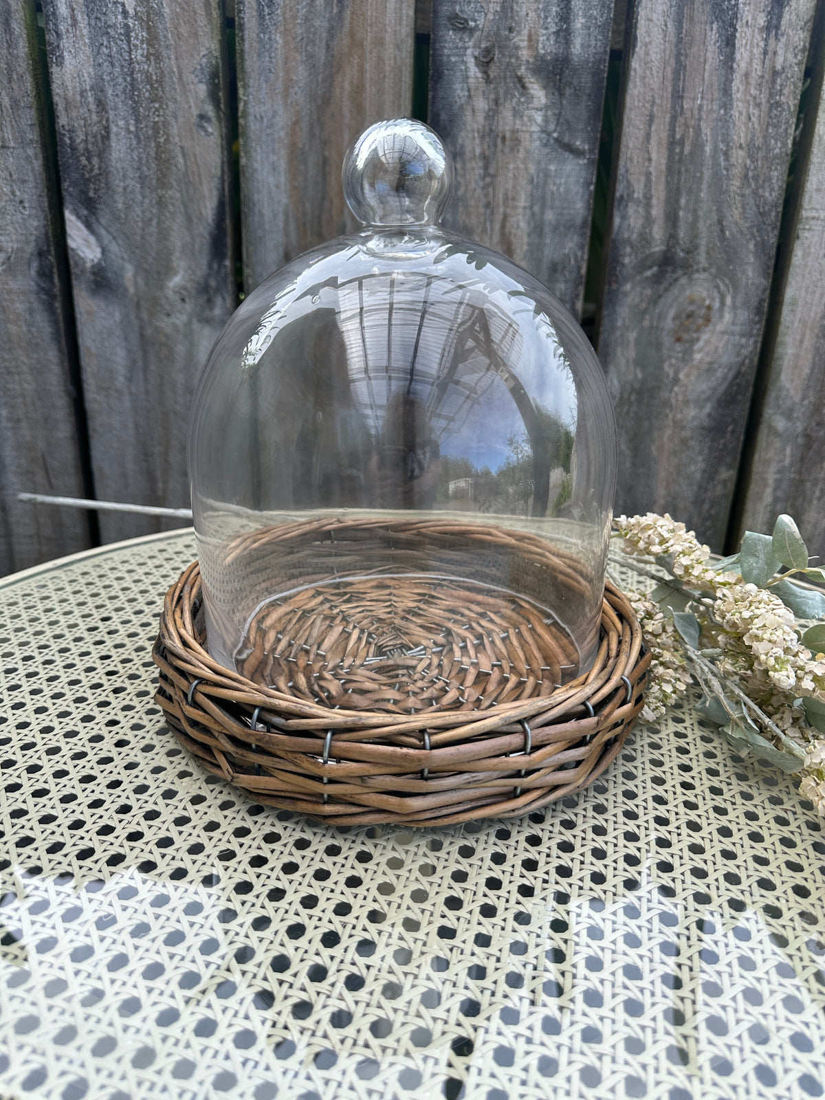 Decorative glass dome with wicker base on a textured surface. Riviera Cloche Dome on Base Small by French Country Collections