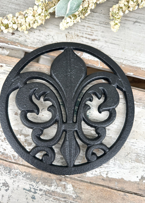 Black cast iron trivet with fleur-de-lis design on a wooden surface.