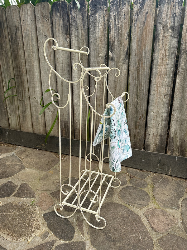 Decorative metal stand with a wooden fence and stone ground in the background. Metal Towel Rail w Shelf Small Aged Cream