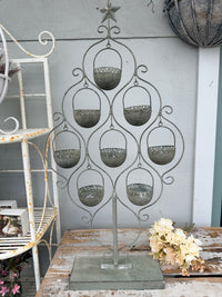 A distressed gray metal candle holder tree with nine hanging candle cups, displayed on a rustic wooden table against a gray wall.
