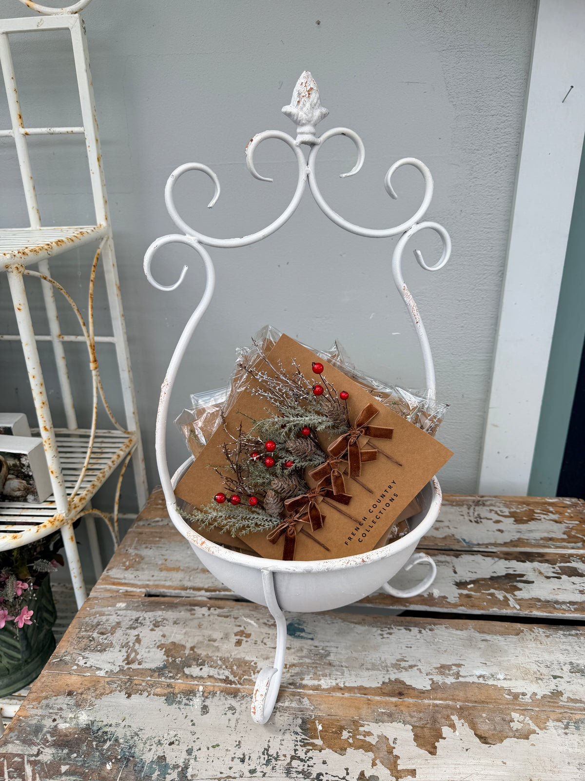 A white distressed metal bowl basket with an ornate scroll frame, holding a brown kraft gift box tied with a ribbon and decorated with evergreen branches and red berries, set on a rustic wooden table against a gray wall.