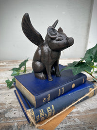 Decorative figurine of a winged pig on top of books with a plant in the background. Flying Pig Black Cast iron