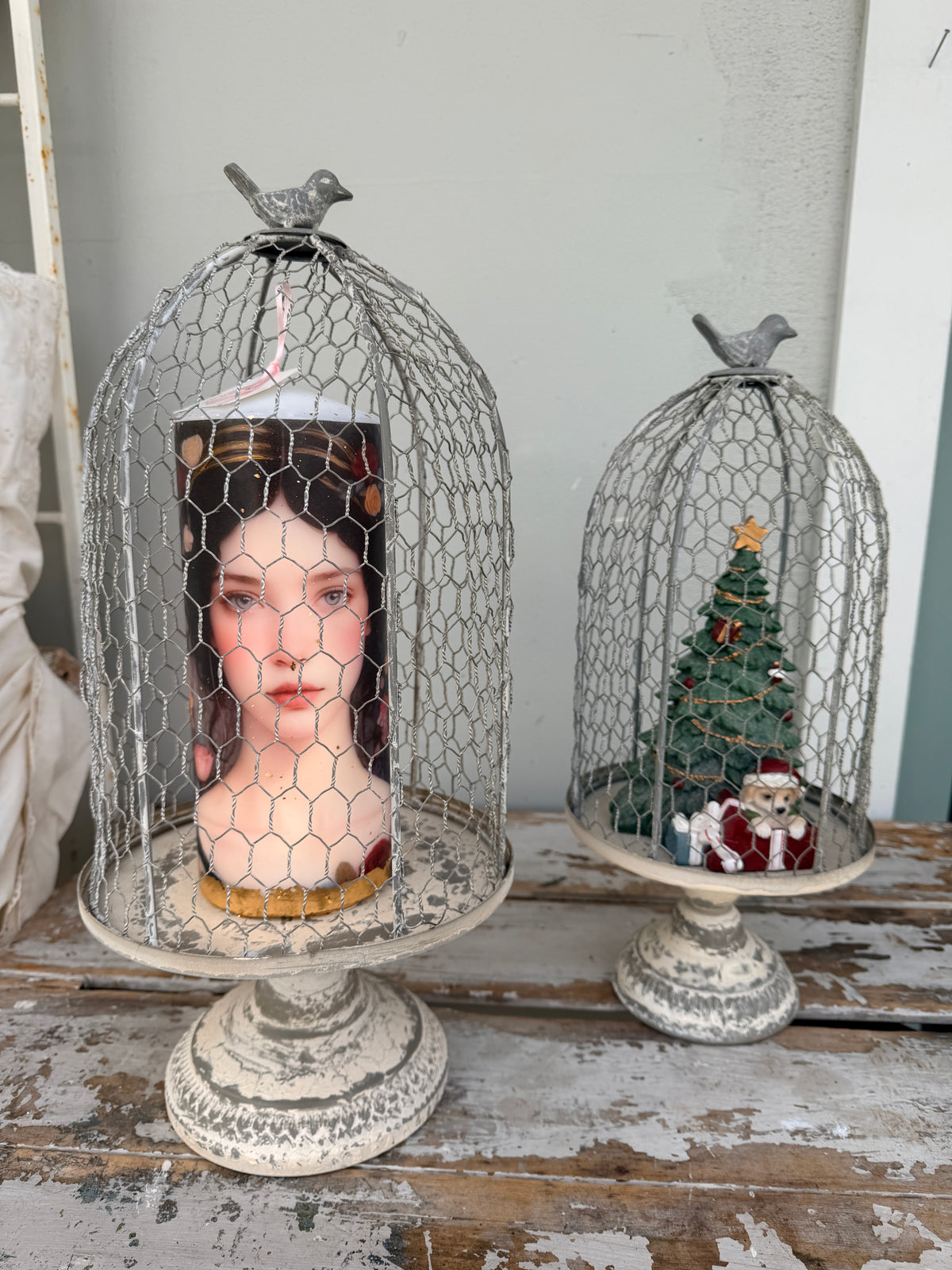 Two distressed white metal birdcage candle holders on pedestals, one holding a portrait candle and the other a miniature Christmas tree with Santa, displayed on a rustic wooden surface.
