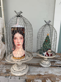 Two distressed white metal birdcage candle holders on pedestals, one holding a portrait candle and the other a miniature Christmas tree with Santa, displayed on a rustic wooden surface.