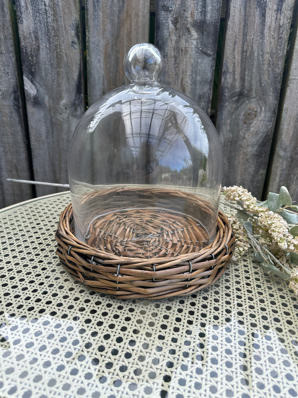 Decorative glass dome with wicker base on a textured surface. Riviera Cloche Dome on Base Small by French Country Collections 