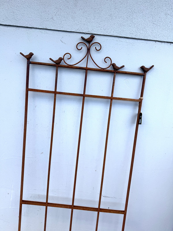 A rusted brown metal garden trellis with decorative bird perches at the top corners and scrollwork, shown leaning against a white stucco wall.