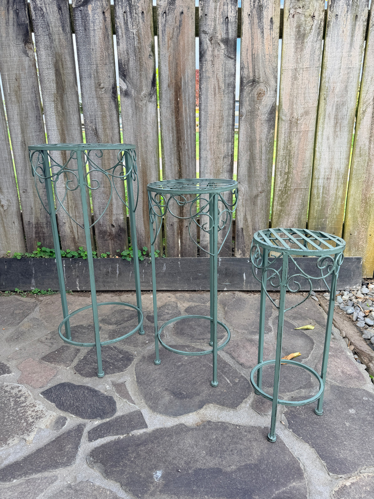 Three green metal garden plant stands of different heights arranged on a stone patio in front of a wooden fence.
