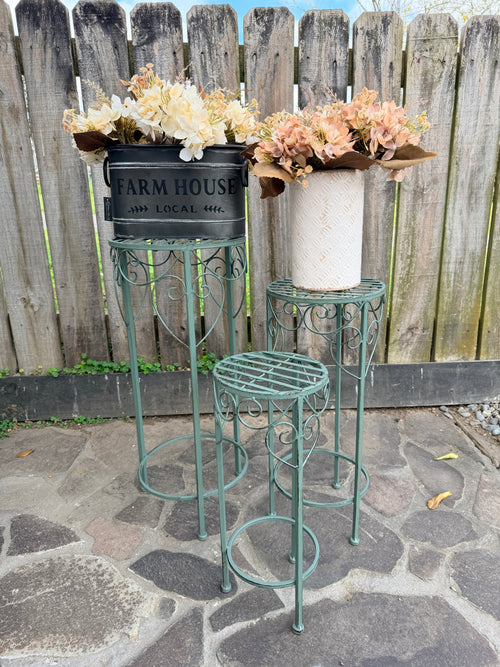 Three metal garden plant stands in a distressed teal green finish, styled with faux flowers in rustic containers in front of a weathered wooden fence on a stone patio.