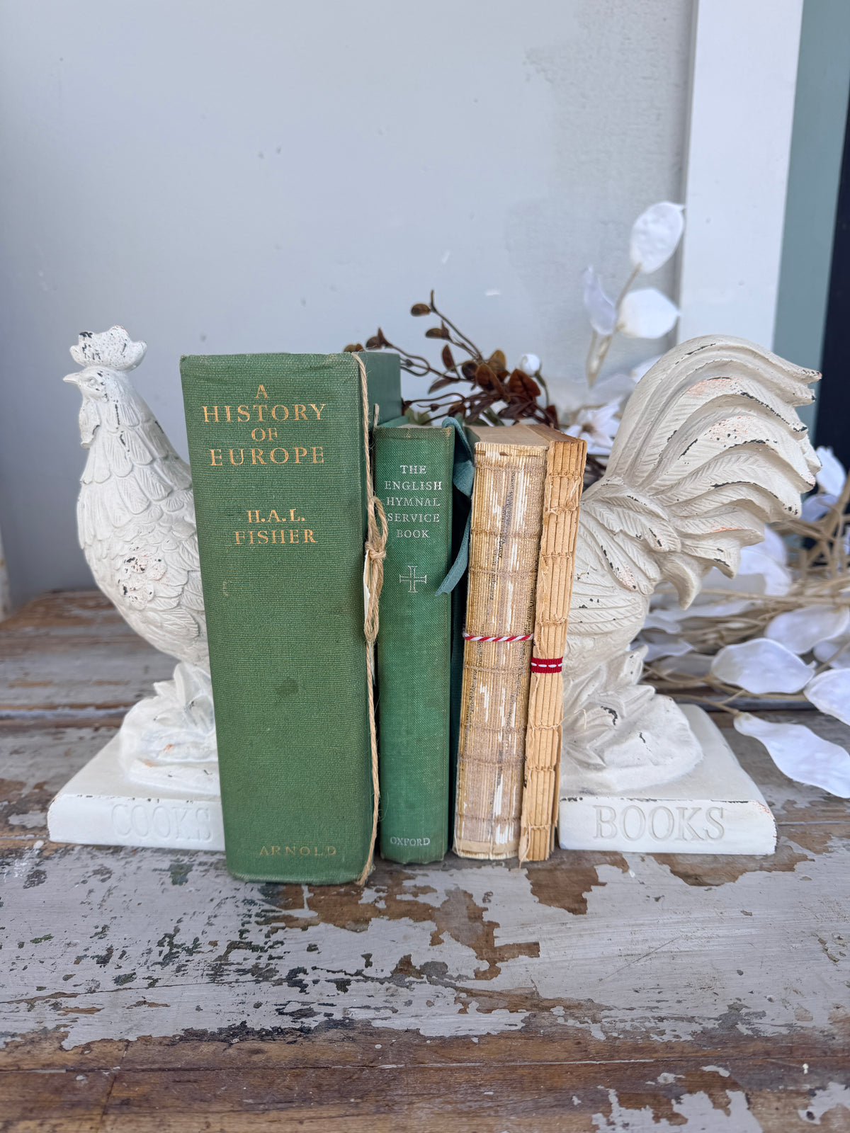 A pair of distressed white rooster bookends holding two vintage-style tan books tied with red string, French Rooster white Book Ends set of 2