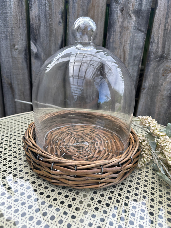 Glass dome with wicker basket on a textured surface. Riviera Cloche Dome on Base Large by French Country Collections
