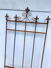 A rusted brown wrought iron decorative gate screen with five fleur-de-lis finials, shown leaning against a light gray textured wall.