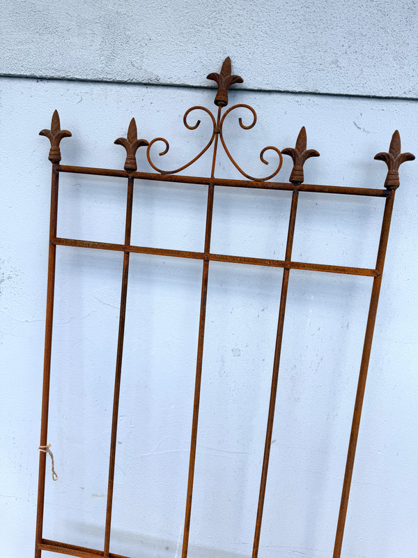 A rusted brown wrought iron decorative gate screen with five fleur-de-lis finials, shown leaning against a light gray textured wall.