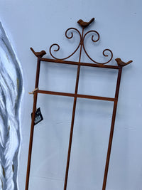 A rusted brown metal garden trellis with decorative bird perches at the top corners, shown leaning against a light blue wall.