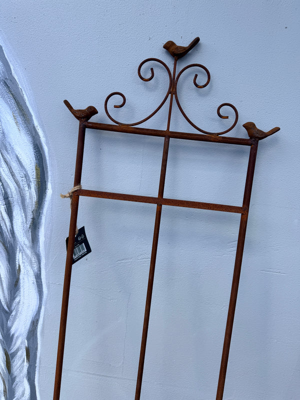A rusted brown metal garden trellis with decorative bird perches at the top corners, shown leaning against a light blue wall.
