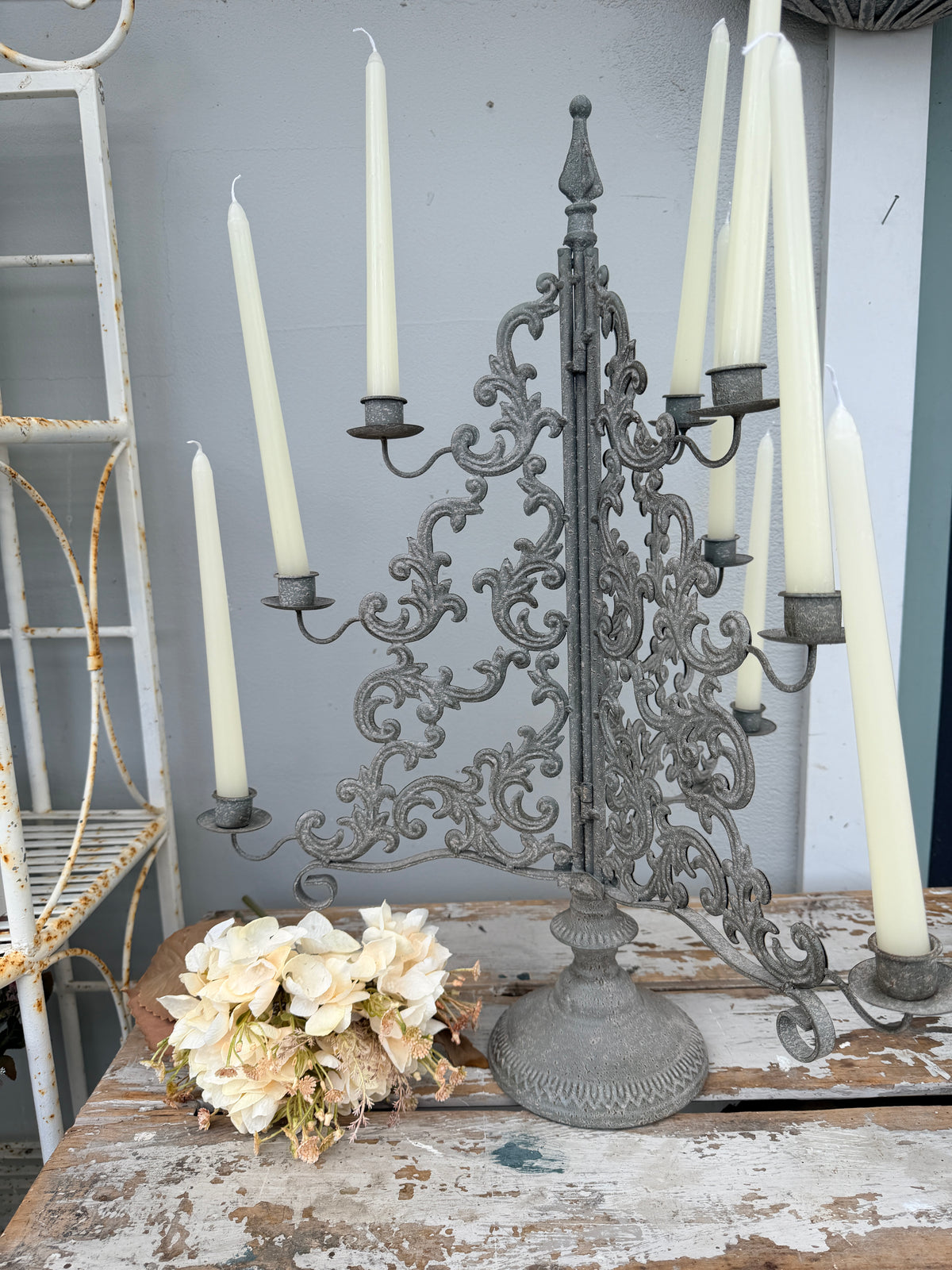 An ornate vintage-style cast iron candelabra with scrollwork and multiple arms, holding cream taper candles, displayed on a rustic wooden table next to a bouquet of cream flowers.