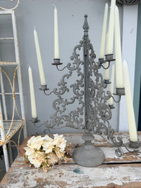 An ornate vintage-style cast iron candelabra with scrollwork and multiple arms, holding cream taper candles, displayed on a rustic wooden table next to a bouquet of cream flowers.