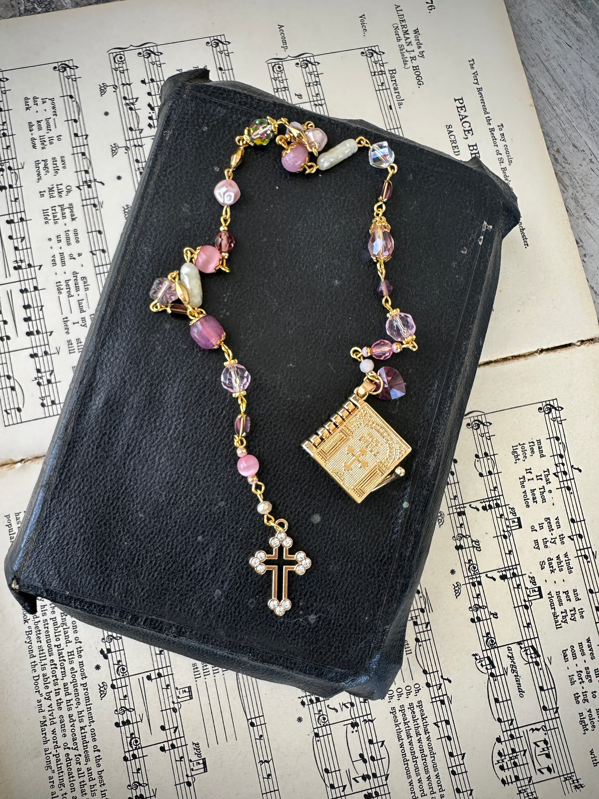 Rosary with colorful beads and a gold cross on a black leather book with music sheet background. Decor prayer beads Pink w Lords Prayer book