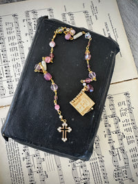 Rosary with colorful beads and a gold cross on a black leather book with music sheet background. Decor prayer beads Pink w Lords Prayer book