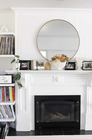 White fireplace with a round mirror above it, decorated with books and plants. crisp white chalk paint