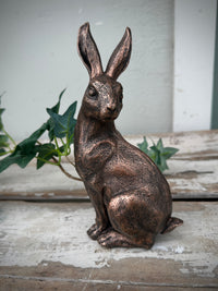 Bronze rabbit statue on a rustic wooden surface with greenery in the background. Bronze Rabbit Sitting head turned