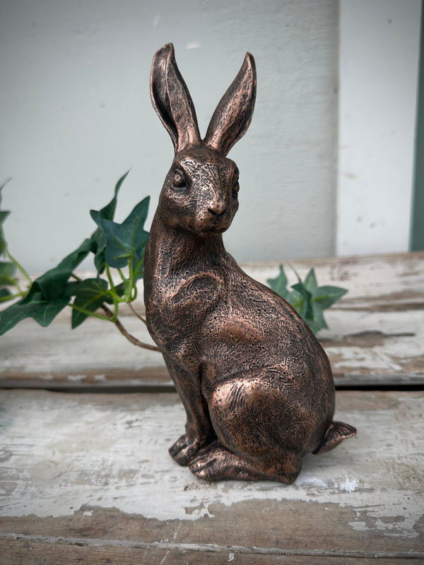 Bronze rabbit statue on a rustic wooden surface with greenery in the background. Bronze Rabbit Sitting head turned