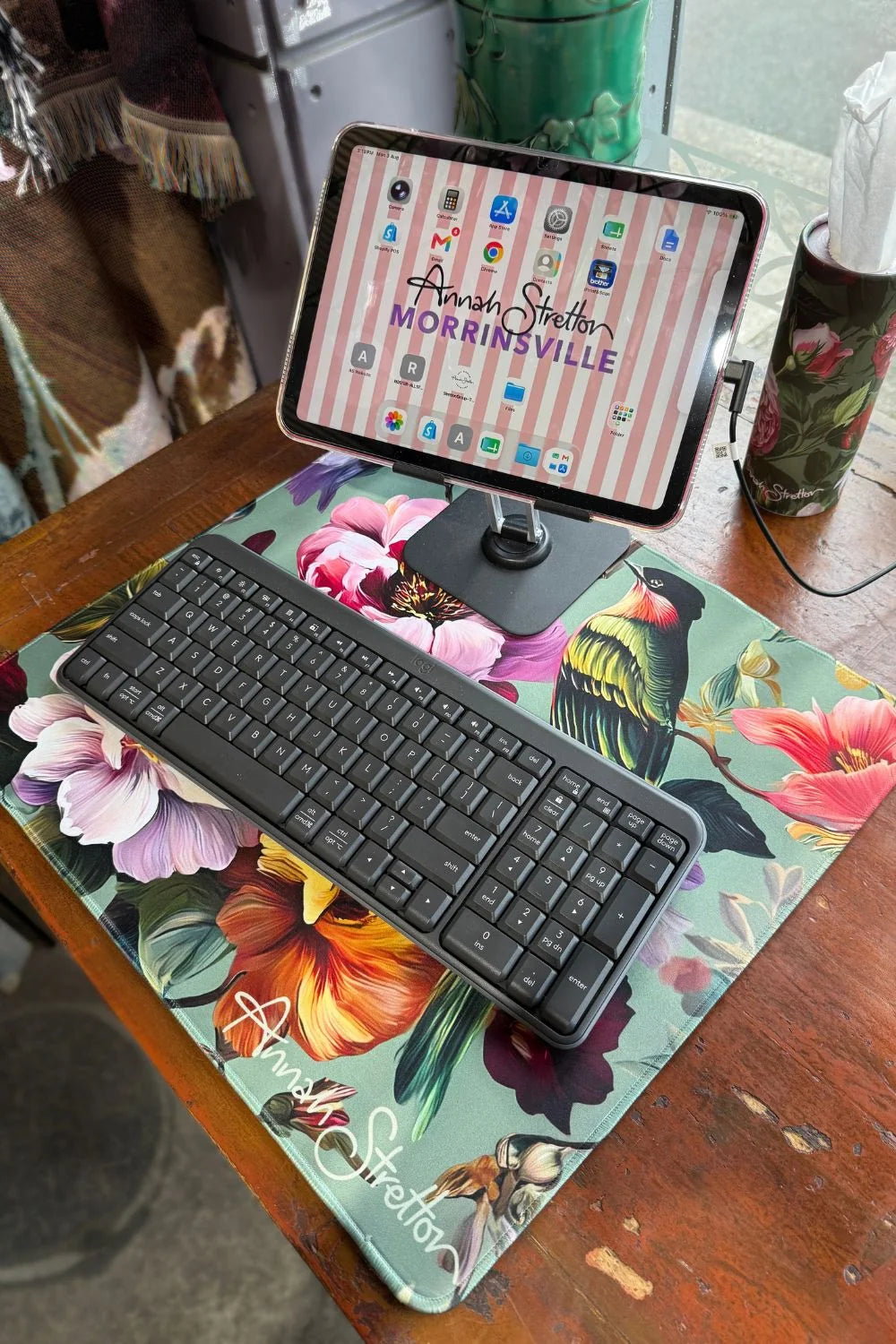 A floral desk mat with a black wireless keyboard and an iPad on a stand, placed on a wooden table in a home setting. Peony Aviary Computer Desk Mat small Annah Stretton
