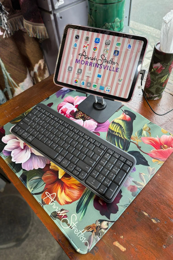 A floral desk mat with a black wireless keyboard and an iPad on a stand, placed on a wooden table in a home setting. Peony Aviary Computer Desk Mat small Annah Stretton