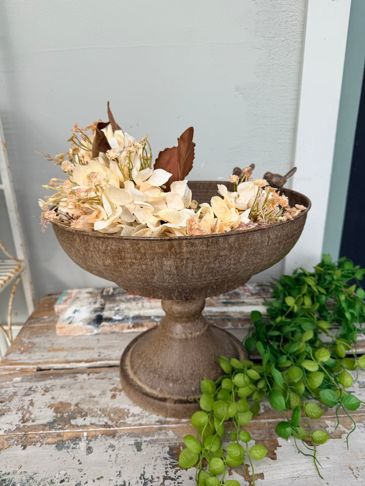 A distressed brown pedestal bowl filled with dried cream flowers and leaves, styled on a rustic white wooden table with green trailing vines in the foreground.