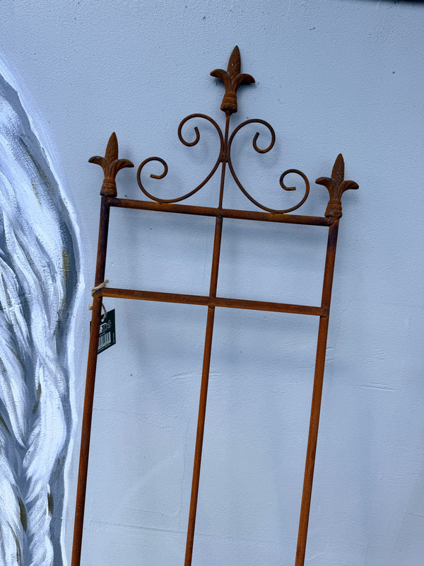 A rusted brown metal garden stake or trellis frame with decorative scrollwork and fleur-de-lis tops, leaning against a light blue wall.