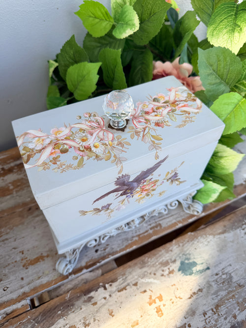 Decorative box with floral and bird design on a stand with greenery in the background. Filigree Blue Lidded Box