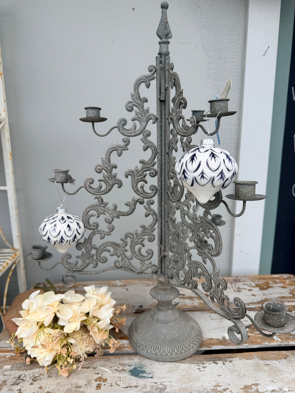 An ornate distressed gray cast iron candelabra with multiple candle holders and two hanging white ceramic ornaments decorated with blue patterns, displayed on a rustic wooden surface.