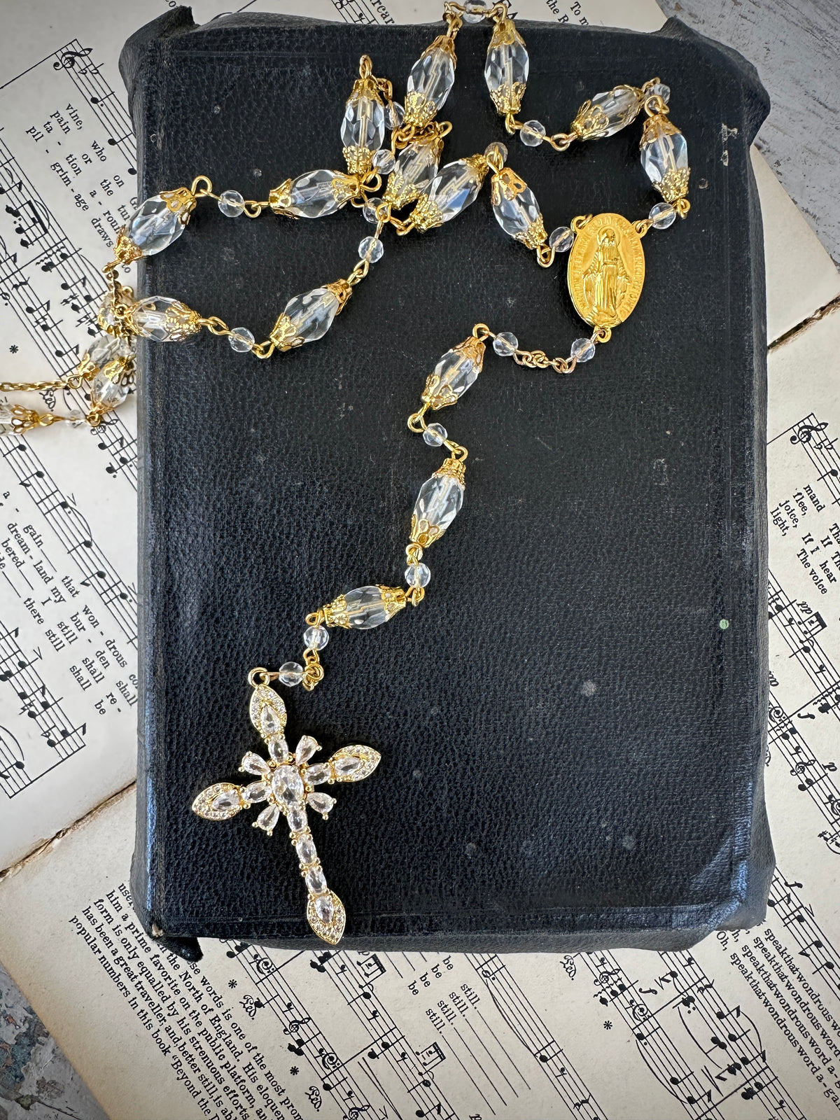Rosary with clear beads and gold accents on a black leather book with music sheet background. Decor prayer beads longer in clear and gold