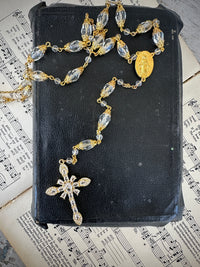 Rosary with clear beads and gold accents on a black leather book with music sheet background. Decor prayer beads longer in clear and gold