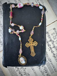 Rosary with pink and gold beads and cross on a black book with music sheet background. Decor prayer beads Pinks w large cross