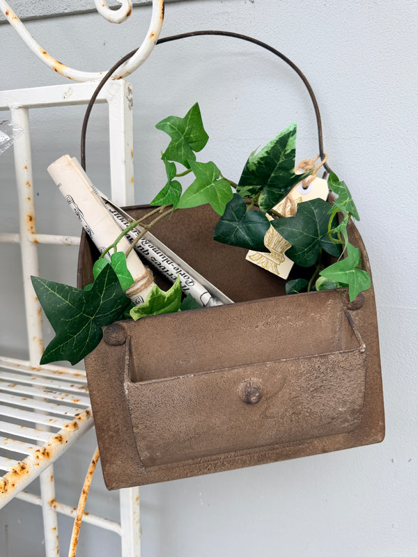 A rustic brown metal wall basket with a curved handle, styled with green ivy leaves and a rolled newspaper,