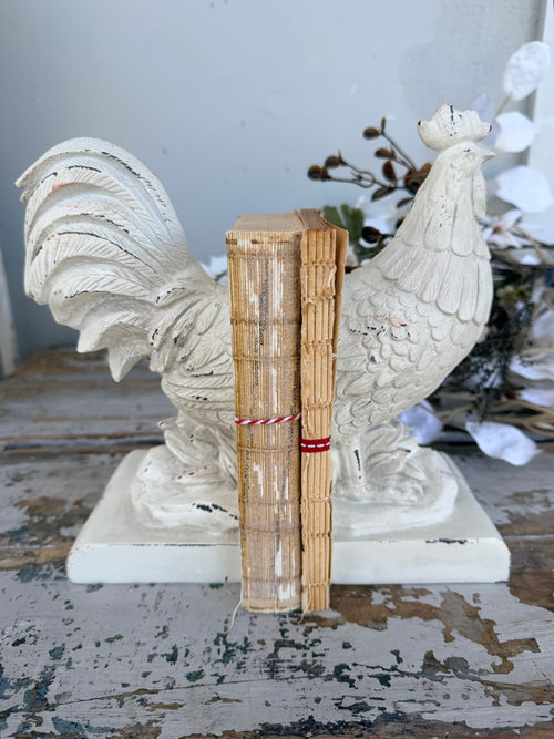 A pair of distressed white rooster bookends holding two vintage-style tan books tied with red string, French Rooster white Book Ends set of 2
