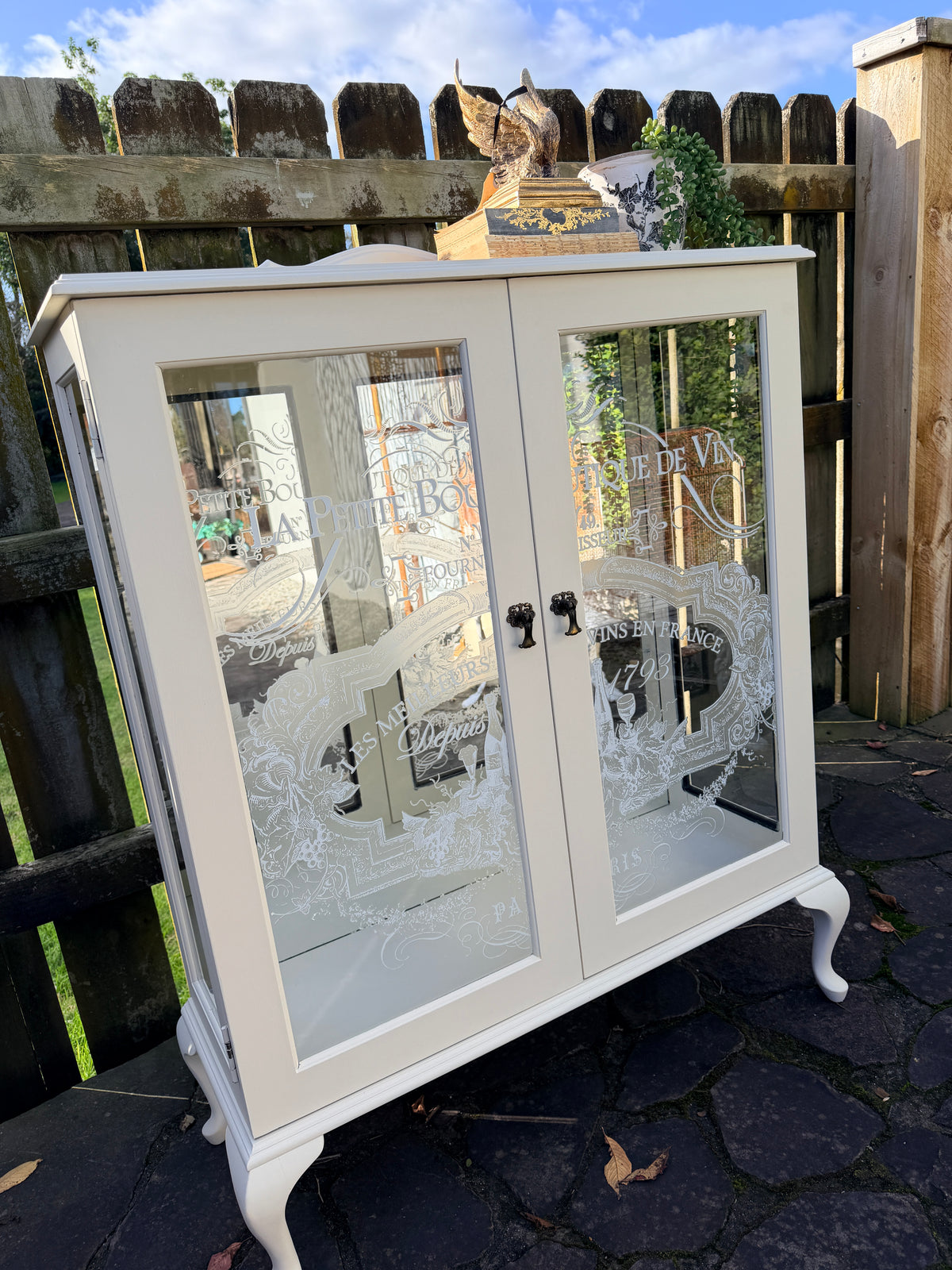 Cabinet with glass doors featuring embossed designs and text. Limestone Cream & White mirrored Display Cabinet with glass shelves