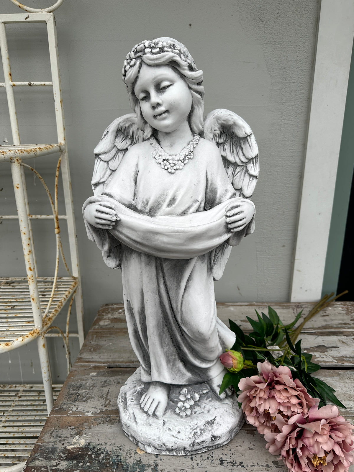 Statue of an angel with wings spread on a wooden surface with flowers. Girl Angel Statue with swag 