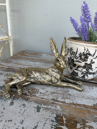Decorative gold rabbit figurine on a rustic wooden surface with a pot of purple flowers in the background. Golden Rabbit lying down