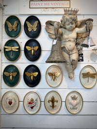 A collection of decorative wall-mounted art pieces, including oval framed butterflies, dragonflies, bees, cherub figurines, and decorative plaques with hearts, fleur-de-lis. A sign at the top reads 'PAINT ME VINTAGE'.