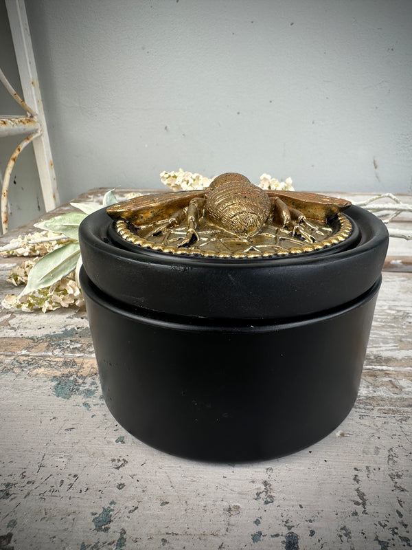 Black container with a gold lid featuring a design on a textured bee surface