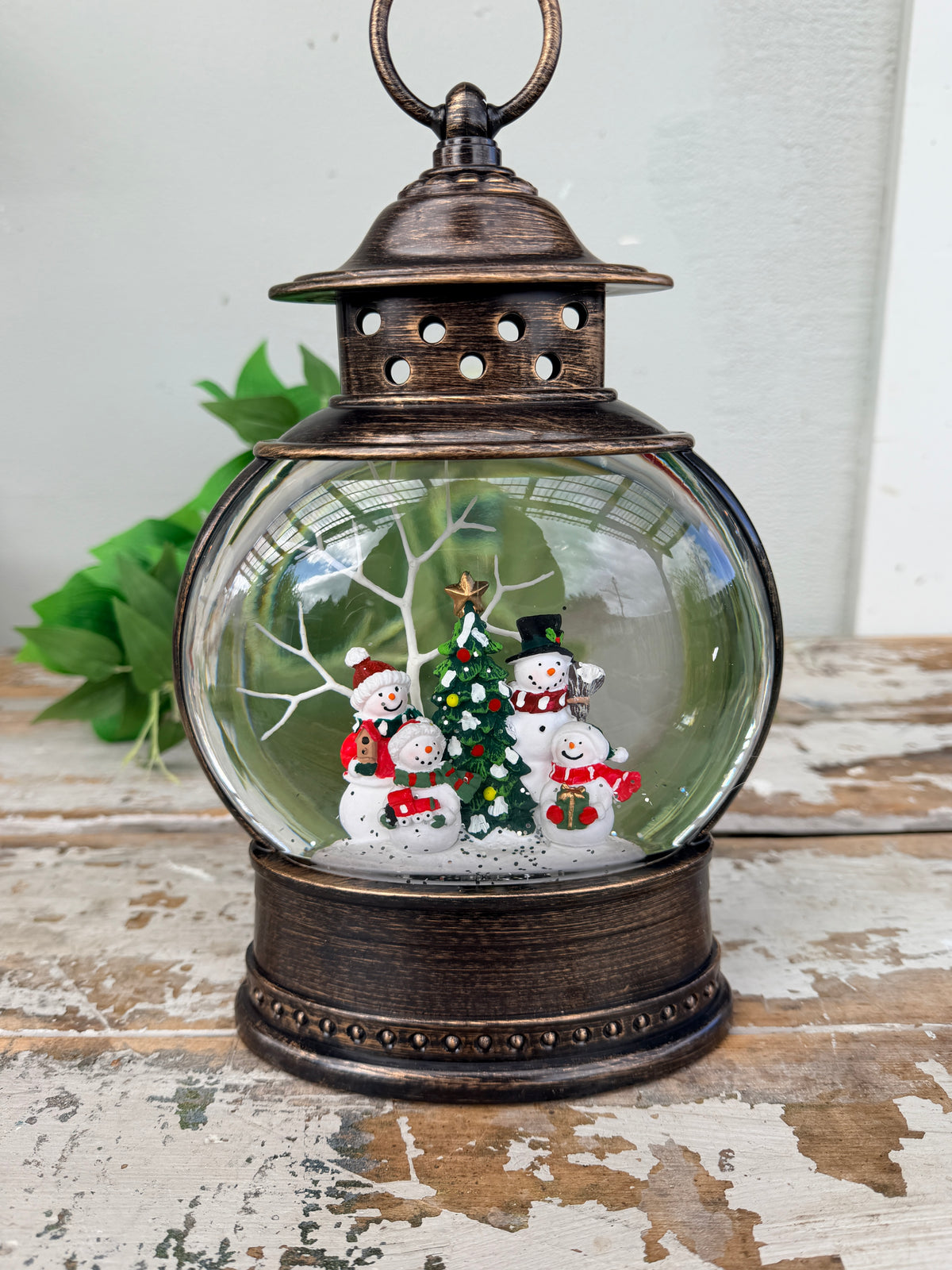 A decorative glass snow globe lantern with an antique bronze metal frame and handle, featuring a small Christmas tree and snowman figures inside, displayed on a rustic wooden surface.