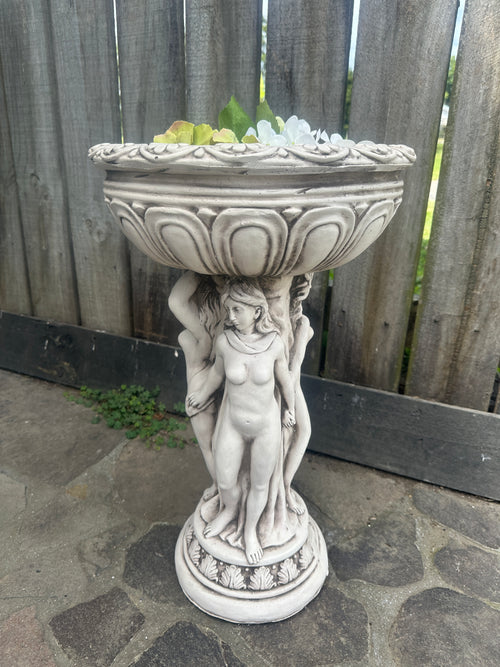 Decorative stone statue on a pedestal with intricate details, set against a wooden fence and stone wall. Goddess Statue