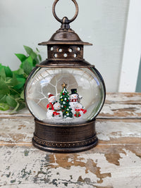 A rustic bronze lantern snow globe with a clear glass dome featuring a small Christmas tree and snowman scene, displayed on a distressed wooden surface.