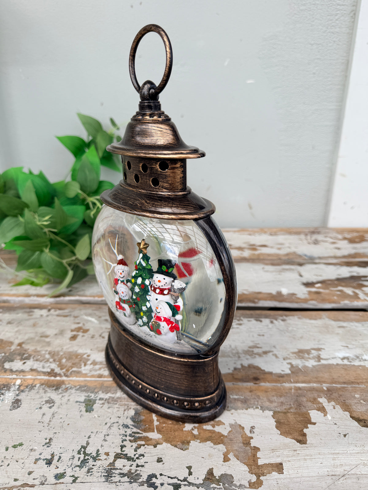A vintage-style metal lantern with a clear glass globe containing painted snowmen and a Christmas tree, displayed on a distressed wooden surface against a light wall with green leaves in the background.