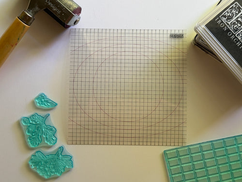A clear acrylic gridded stamp mat with circular guide lines, styled on a white surface with craft tools including a rubber stamp, ink pad, and green rubber stamps. IOD Mini Thin Stamp Mount