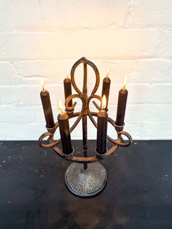 A five-arm wrought iron candelabra with a distressed dark brown finish, holding five dark brown taper candles that are lit, set on a dark surface against a white brick wall.
